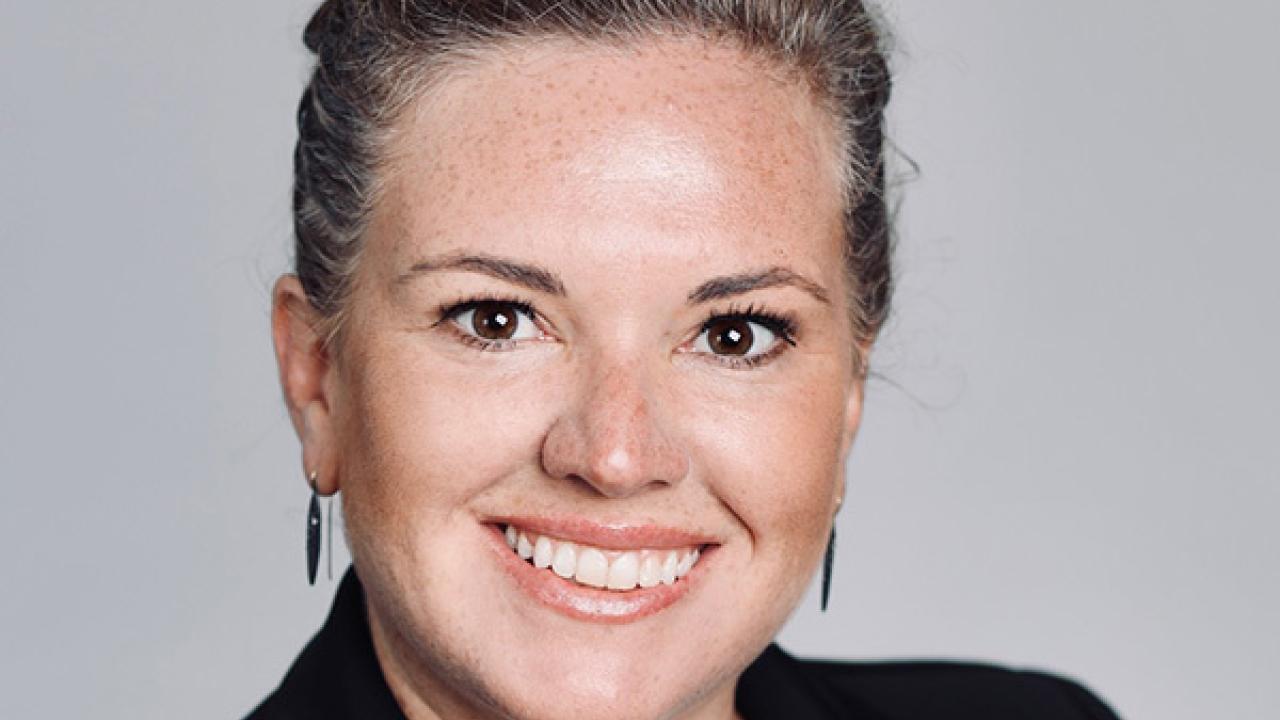 Professional headshot of smiling blonde woman with bun, black blazer, gray background